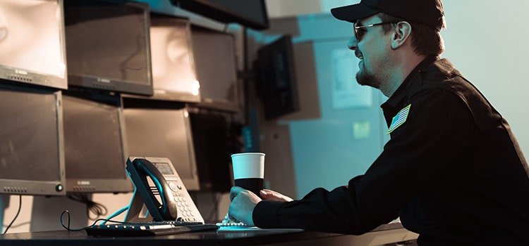 guard sits at desk with coffee and phone watching several monitors