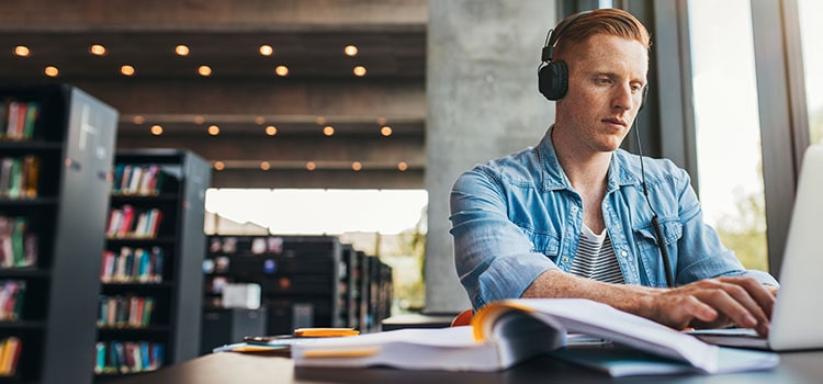student wearing headphones types on laptop seated at textbook covered desk in library