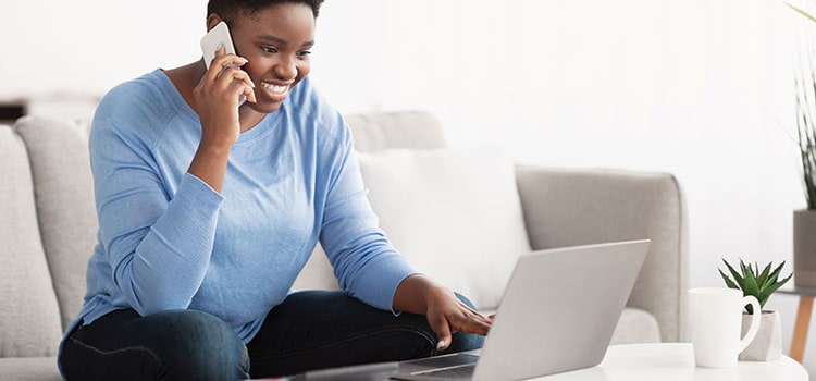 person on couch talks on phone while smiling at laptop screen